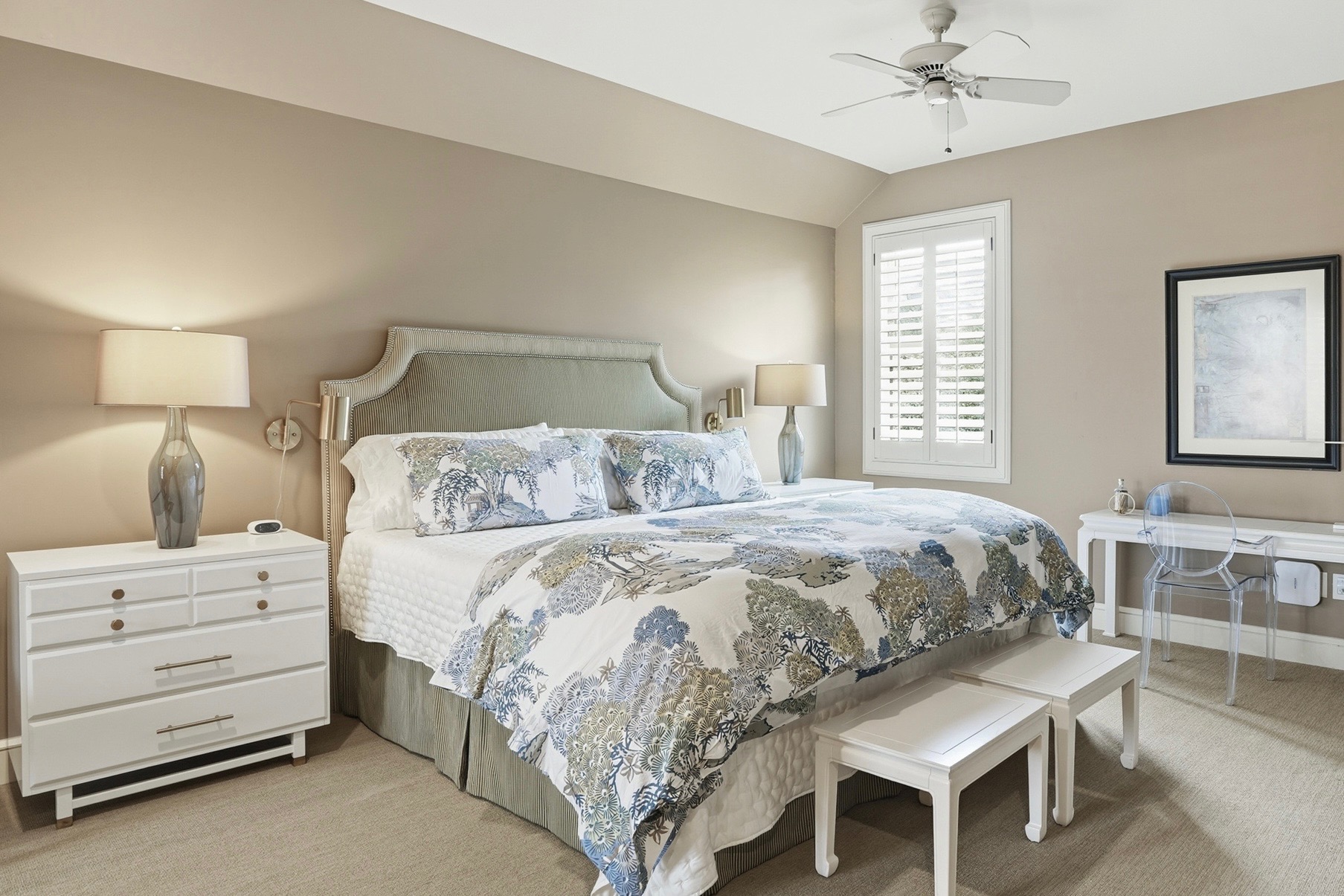 Serene guest bedroom with upholstered headboard, botanical bedding, plantation shutters, and a ghost-chair writing desk